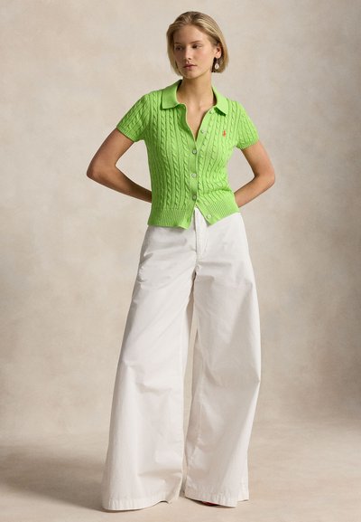 Nainen, jolla on kirkkaanvihreä, lyhythihainen palmikkoneuletakki ja leveälahkeiset valkoiset housut, seisoo käsivarret selän takana.