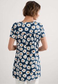 Gėlių raštuota tunika su tamsiai mėlynu fonu, puošta baltomis ramunėlėmis su oranžinėmis šerdimis. Pagaminta iš mink�štos medžiagos, turi apvalų iškirpimą ir trumpas rankoves.