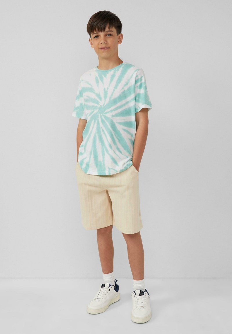 Tie-dye T-shirt met korte mouwen in turquoise en wit, gecombineerd met lichtgele gestreepte shorts. Witte sneakers maken de outfit compleet.