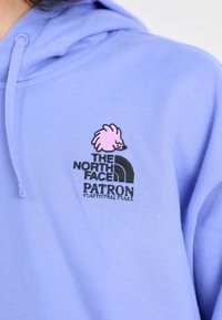 Ljuslila hoodie med en rosa igelkottslogotyp och texten "The North Face", med svart "PATRON" och slogan i kontrasterande färger.