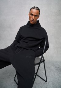 Sudadera negra con alto cuello y joggers ajustados a juego, con un logo bordado en la pierna del pantalón, sentado en una silla de metal.