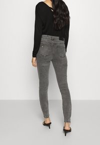 Jeans skinny de talle alto en gris con un acabado desgastado, tres bolsillos traseros y un detalle de logo en la cinturilla negra, combinados con una blusa negra y tacones.
