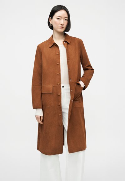 Γυναίκα που φοράει καφέ μακρύ παλτό με κουμπιά και τσέπες, πάνω από λευκό τοπ και λευκό παντελόνι με φαρδιά πόδια, στέκεται μπροστά σε ένα απλό φόντο.