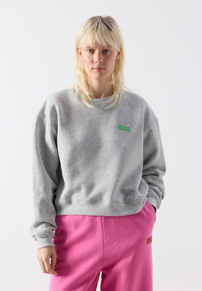 Blond woman wearing a gray sweatshirt and pink sweatpants with green "American Vintage" logos on each garment, standing against a white background.
