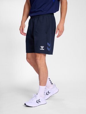 Mann trägt marineblaue Sportshorts und weiße Sportschuhe mit passenden weißen Socken vor einfarbigem Hintergrund.