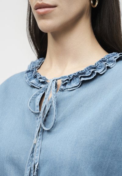 Κοντινό πλάνο γυναίκας που φοράει πουκάμισο με ανοιχτό μπλε γιακά με βολάν δεμένο με φιόγκο, με ορατά χρυσά σκουλαρίκια σε σχήμα κρίκου.