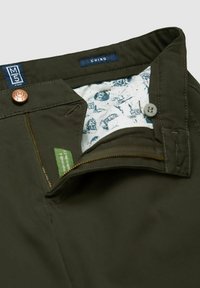 Donkergroene chino broek met een rits, koperen knoopsluiting en een gebloemde binnenvoering met blauwe illustraties. Gelabeld "M5 CHINO."