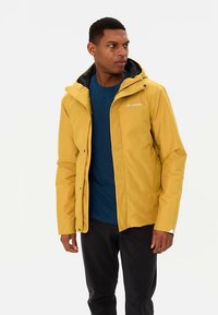 Geel waterdicht jack met capuchon, voorzien van een ritssluiting aan de voorkant, zwarte accenten en een logo. Draagt over een blauw shirt en zwarte broek.