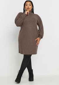 Brun ribbad stickad klänning med hög krage, långa ballongärmar och rak silhuett. Bärs med svarta ogenomskinliga strumpbyxor och ankelstövlar.