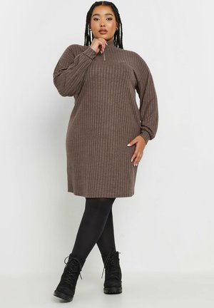 Robe en tricot côtelé marron avec un col montant, des manches ballons longues et une silhouette droite. Portée avec des collants opaques noirs et des bottines.