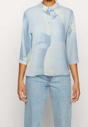 Kobieta w jasnoniebieskiej zapinanej na guziki bluzce z abstrakcyjnymi wzorami i rękawami do trzy czwarte, w połączeniu z jasnoniebieskimi dżinsami.