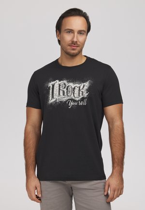 Dorosły mężczyzna o ciemnych włosach, ubrany w czarną koszulkę z białym napisem "I ROCK your roll" na jednolitym tle.