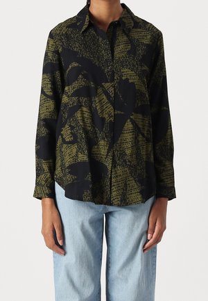 Personne portant une chemise à manches longues boutonnée avec des motifs abstraits de feuilles noires et vert olive, et un jean bleu clair.