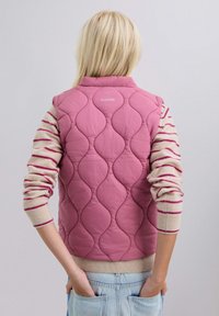 Chaleco acolchado rosa con un patrón de olas, con cuello alto y el nombre de la marca "SCALPERS" en la parte de atrás. Lleva puesto un suéter de punto a rayas.