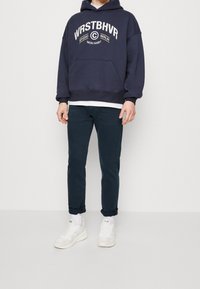 Sudadera navy con texto blanco, bolsillo frontal y puños acanalados, combinada con pantalones ajustados de color verde oscuro y zapatillas blancas.