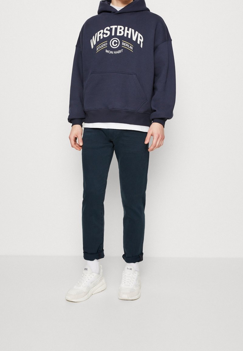 Sudadera navy con texto blanco, bolsillo frontal y puños acanalados, combinada con pantalones ajustados de color verde oscuro y zapatillas blancas.