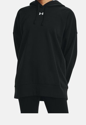 Osoba nosząca czarną, dużą bluzę z kapturem z białym logo Under Armour na piersi oraz czarne legginsy na jednolitym tle.