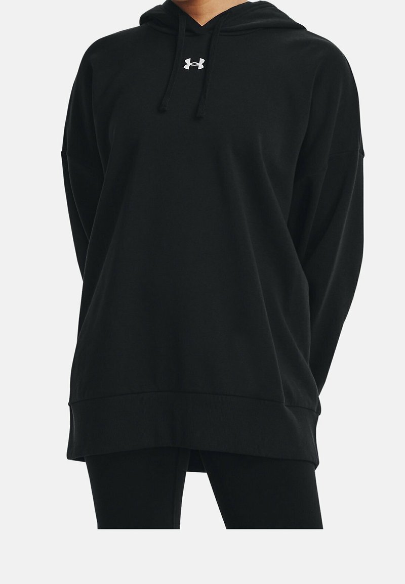 Personne portant un sweat à capuche noir oversize avec un logo Under Armour blanc sur la poitrine et des leggings noirs devant un fond uni.