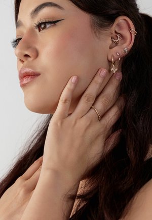 Junge Frau mit mehreren goldenen Ohrringen, einem goldenen Ring, Flügel-Eyeliner und langen dunklen Haaren, die ihren Hals und die Kieferlinie berührt.