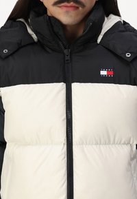 Chaqueta acolchada con diseño dividido; parte superior en negro, parte inferior en blanco. Presenta un cuello alto, cierre de cremallera y un pequeño logo en el pecho.