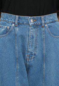 Jean en denim avec bouton et fermeture éclair à l'avant, couture orange et une chemise noire rentrée dans la taille.