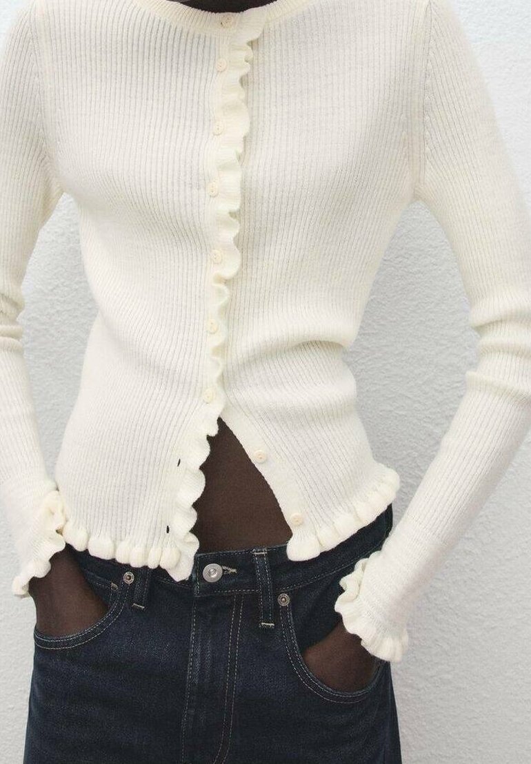 Personne portant un cardigan côtelé crème avec des bords froncés et un pantalon noir, mains dans les poches, sur un fond clair uni.