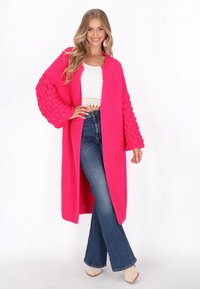 Růžový pletený kardigan s texturovanými, oversized rukávy, nosí se přes bílý crop top a rozšířené modré džíny, s dlouhým otevřeným designem.