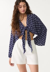 Crop top bleu marine avec un nœud à l'avant, orné d'un motif floral blanc répété et de manches amples. Associé à un short blanc taille haute.