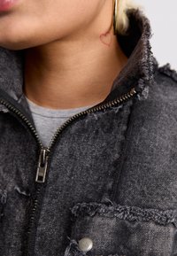 Σκούρο γκρι τζιν μπουφάν με ξέφτια στις άκρες, φερμουάρ μπροστά και τσέπη με κουμπί που κλείνει με κλιπ. Διαθέτει ήπια υφή και μεταλλικό φερμουάρ.