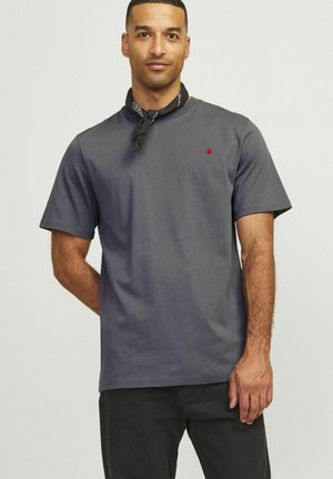 Graues Baumwoll-T-Shirt mit kurzen Ärmeln, versehen mit einem kleinen roten Logo auf der Brust. Das Model trägt ein schwarzes Bandana um den Hals.