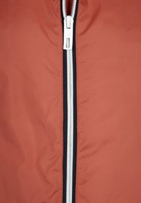 Veste légère orange avec une fermeture éclair à l'avant. Comprend une fermeture éclair blanche entourée de détails en tissu noir. Texture lisse et mate.