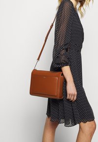 Brun läder crossbody-väska med rektangulär form, minimal hårdvara och en slät yta. Har en avtagbar rem och präglad logotyp.