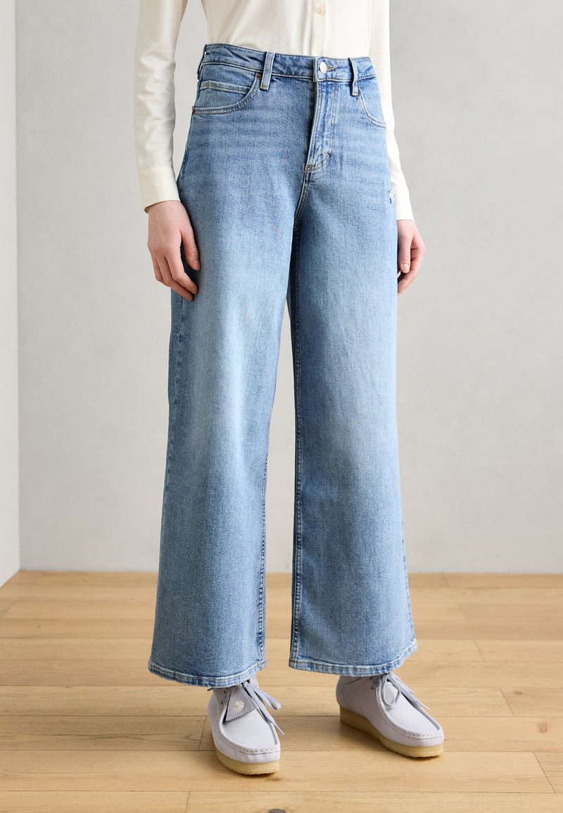 Bredde ben jeans i lys blå denim, med høy midje, frontlommer og rå kant, kombinert med grå semsket skinnsko.