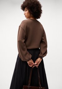 Barna, oversized pulóver puffos ujjakkal, fekete redőzött szoknyával párosítva. A modell egy sima textúrájú barna kézitáskát tart.