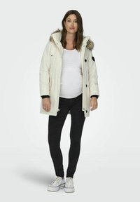 Parka bianca con cappuccio foderato in pelliccia, dettagli neri, tasche frontali e chiusa con bottoni. Indossata sopra una maglietta bianca aderente e jeans neri.
