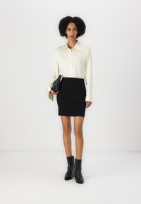 Bluză bej deschis cu guler, fustă mini neagră cu dungi și cizme negre până la gleznă. Modelul ține în mână o geantă mică neagră.