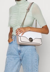 Personne portant un haut froncé vert clair et un jean bleu, tenant un sac à main en cuir beige et blanc avec une bandoulière à chaîne et une fermeture dorée.