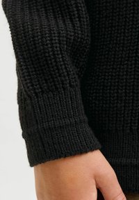 Ärmel eines schwarzen Strickpullis mit einem gerippten Bündchen und einer strukturierten Oberfläche, an der Kante ist eine Hand sichtbar. Weiches, warmes Material.