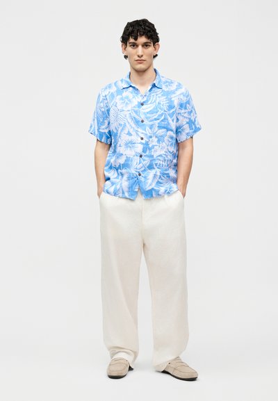 Fiatal férfi, kezét a zsebében tartva, világoskék virágmintás rövid ujjú inget, fehér, bő szárú nadrágot és bézs cipőt visel, fehér háttér előtt.