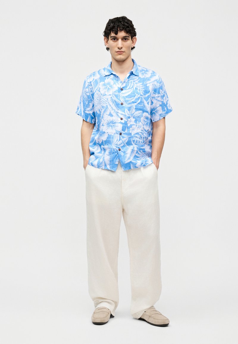 Jonge man staand met handen in broekzakken, gekleed in een lichtblauw bloemenoverhemd met korte mouwen, witte wijde broek en beige schoenen tegen een witte achtergrond.