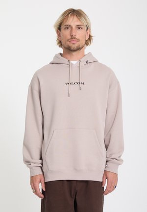 Homme blond aux cheveux courts portant un sweat à capuche beige Volcom et un pantalon marron, debout face à l'avant avec une expression neutre.