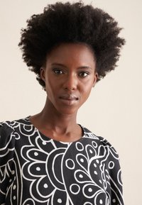 Blusa nera con motivo floreale bianco, caratterizzata da un collo rotondo e maniche arricciate. Tessuto morbido su uno sfondo neutro.