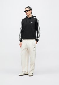 Vyras su pintomis plaukais ir saulės akiniais, vilkintis juodą Adidas džemperį su baltais dryžiais, baltas Adidas kelnes ir baltus sportbačius su juodais dryžiais.