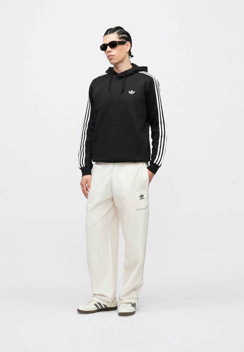 Uomo con capelli intrecciati e occhiali da sole che indossa una felpa nera Adidas con strisce bianche, pantaloni bianchi Adidas e scarpe da ginnastica bianche con strisce nere.