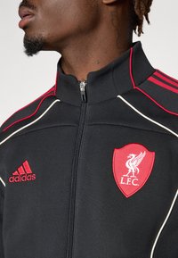 Zwarte sportjas met rode accenten, voorzien van een volledige rits, hoge kraag, geborduurd L.F.C. logo en drie rode strepen op de mouwen.