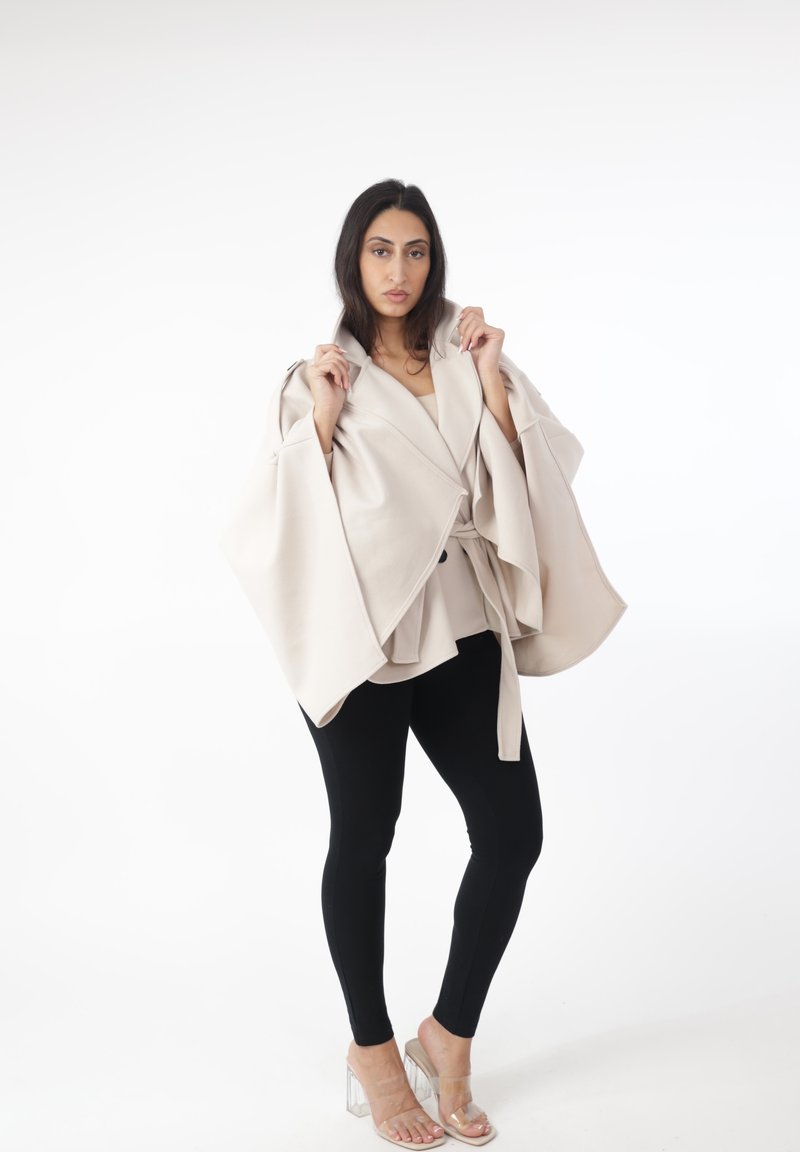 Blazer tipo capa beige y estructurado con mangas anchas, atado a la cintura, usado sobre una camiseta clara y combinado con leggings negros y tacones transparentes.