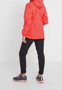 Rode waterdichte jas met een capuchon, elastische mouwen en zijzakken; gecombineerd met zwarte broek en grijze sportschoenen met rode accenten.