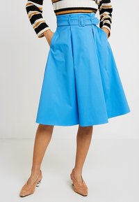 Blå plisserad kjol sydd av ett slätt tyg, med en bred midjeband och ett bälte samt sidofickor. Kombinerad med beige skor.