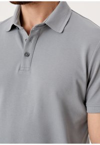 Homme portant un polo gris clair à manches courtes, avec deux boutons fixés, montrant des détails du haut de la poitrine et du col.