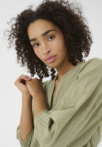 Blouse vert clair, texturée, à coupe ample, avec des manches volumineuses et un décolleté en V, ornée de petits détails en forme de cœur brodés.